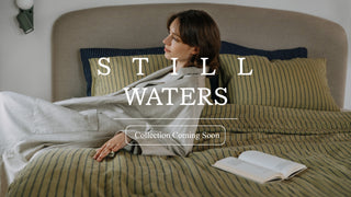 A woman reclines on a bed with striped bedding and pillows in a softly lit bedroom. Large overlaid text reads 'STILL WATERS' with a smaller 'Collection Coming Soon' box below. 