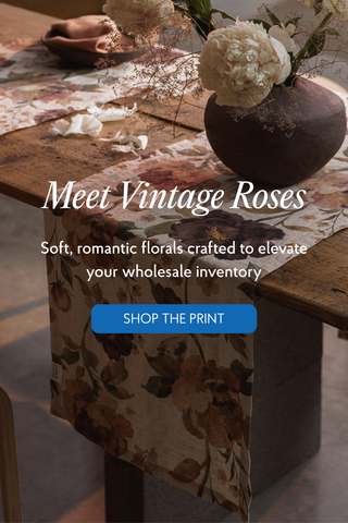 A floral table runner draped over a rustic wooden table, styled with a dark vase of white flowers and overlaid text reading 'Meet Vintage Roses' and 'SHOP THE PRINT'. 